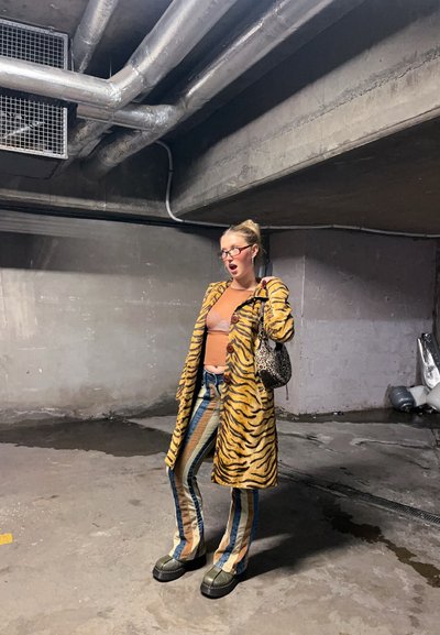 Mujer con pantalones a rayas y abrigo de estampado de tigre posa en un estacionamiento de concreto, sosteniendo un bolso de estampado de leopardo con la mano en la cadera.