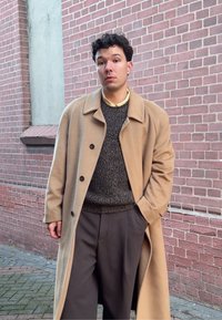 Manteau beige avec col, pull à motifs noirs et gris, et pantalon foncé, sur fond de mur en briques. Accent sur la texture et le contraste des couleurs.
