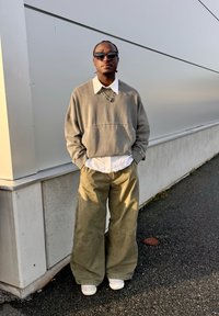 Mann iført store, olivengrønne bukser, grå genser over hvit skjorte, hvite joggesko, solbriller og lagdelte halskjeder som står ved en lysegrå vegg.