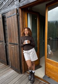 Donna con capelli ricci, vestita con un maglione marrone e pantaloni corti bianchi, tiene una tazza mentre si appoggia a un telaio di porta in legno su un terrazzo all'esterno di una baita.