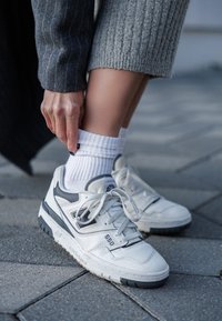 Λευκά αθλητικά παπούτσια με γκρι λεπτομέρειες, τρυπητή επιφάνεια και κορδόνια, φορεμένα με λευκές ριγέ κάλτσες. Η επιφάνεια έχει σχέδιο.