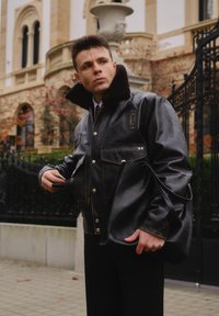 Czarna skórzana kurtka z futrzanym kołnierzem, wyposażona w przednie kieszenie i srebrne guziki, zestawiona z czarną torebką na ramię i dopasowanymi czarnymi spodniami.