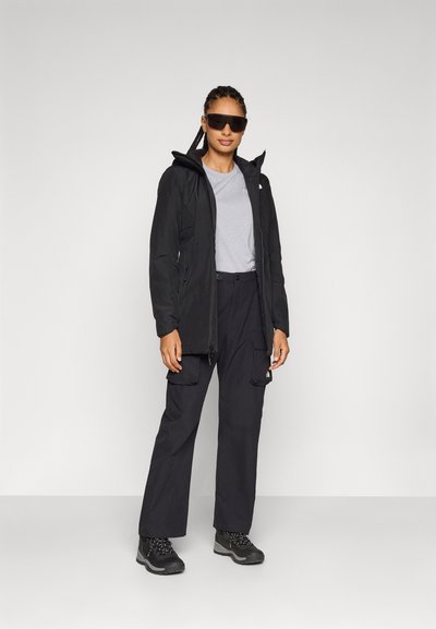 Chaqueta negra impermeable con capucha, camiseta gris, pantalones cargo negros y botas de senderismo negras resistentes, con un diseño moderno y detalles funcionales.