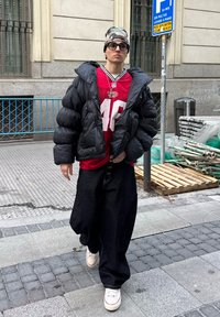 Jeune personne portant une doudoune noire, un maillot de sport rouge, un pantalon noir ample, des baskets blanches, un bonnet camouflage et des lunettes, marchant sur un trottoir urbain.