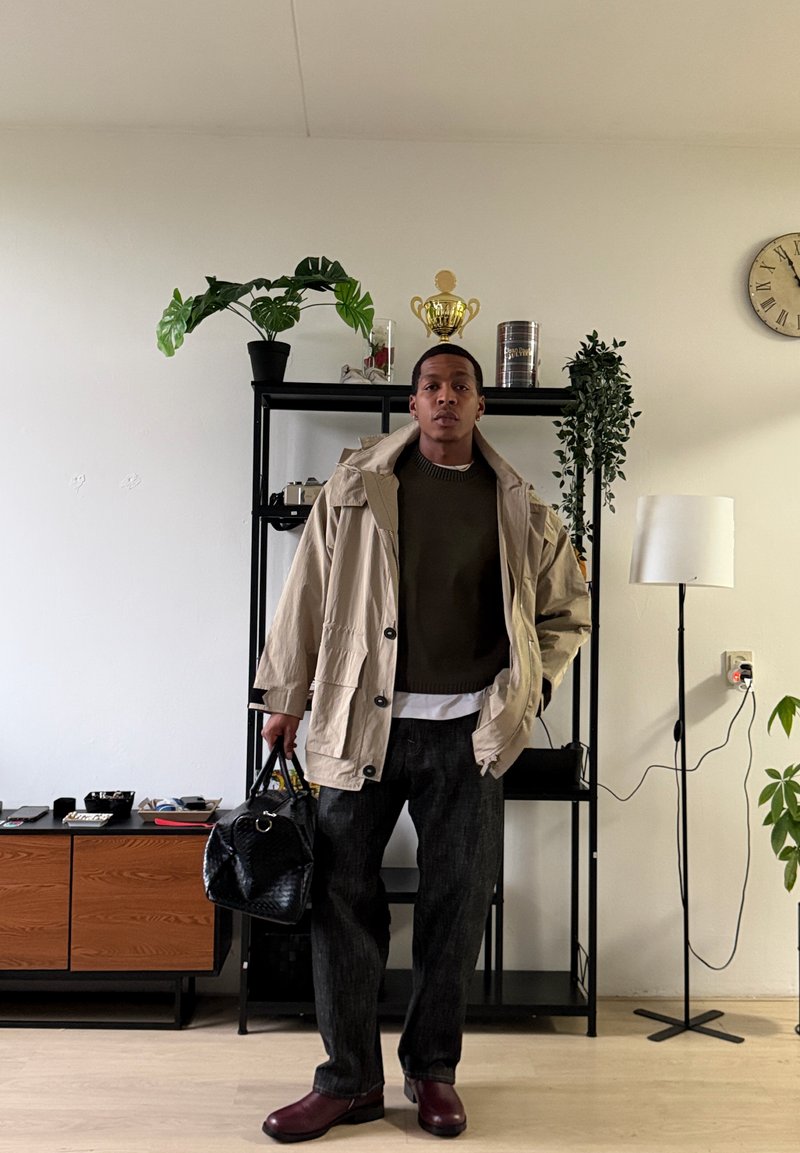 Homme portant une veste beige, un pantalon foncé et des chaussures bordeaux, tenant un sac noir, debout dans une pièce avec des plantes, des étagères, une horloge et une lampe sur pied.