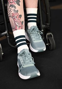 Jambes avec tatouage floral et tatouage de femme, portant des chaussettes blanches à rayures vertes et des chaussures de sport grises, reposant sur les repose-pieds d'un fauteuil roulant.