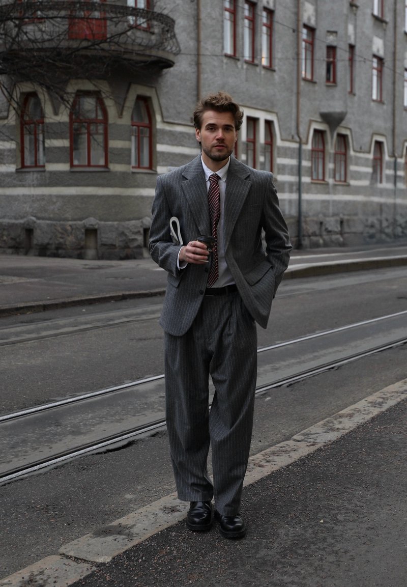 Homme en costume gris à rayures tenant une tasse de café et un journal, marchant dans une rue avec des rails de tramway et un bâtiment en pierre aux fenêtres rouges en arrière-plan.