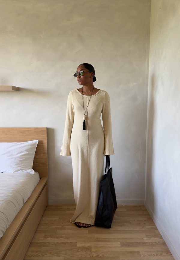 Mujer con vestido largo beige y gafas de sol junto a una cama, sosteniendo un gran bolso negro en una habitación minimalista con suelo de madera.