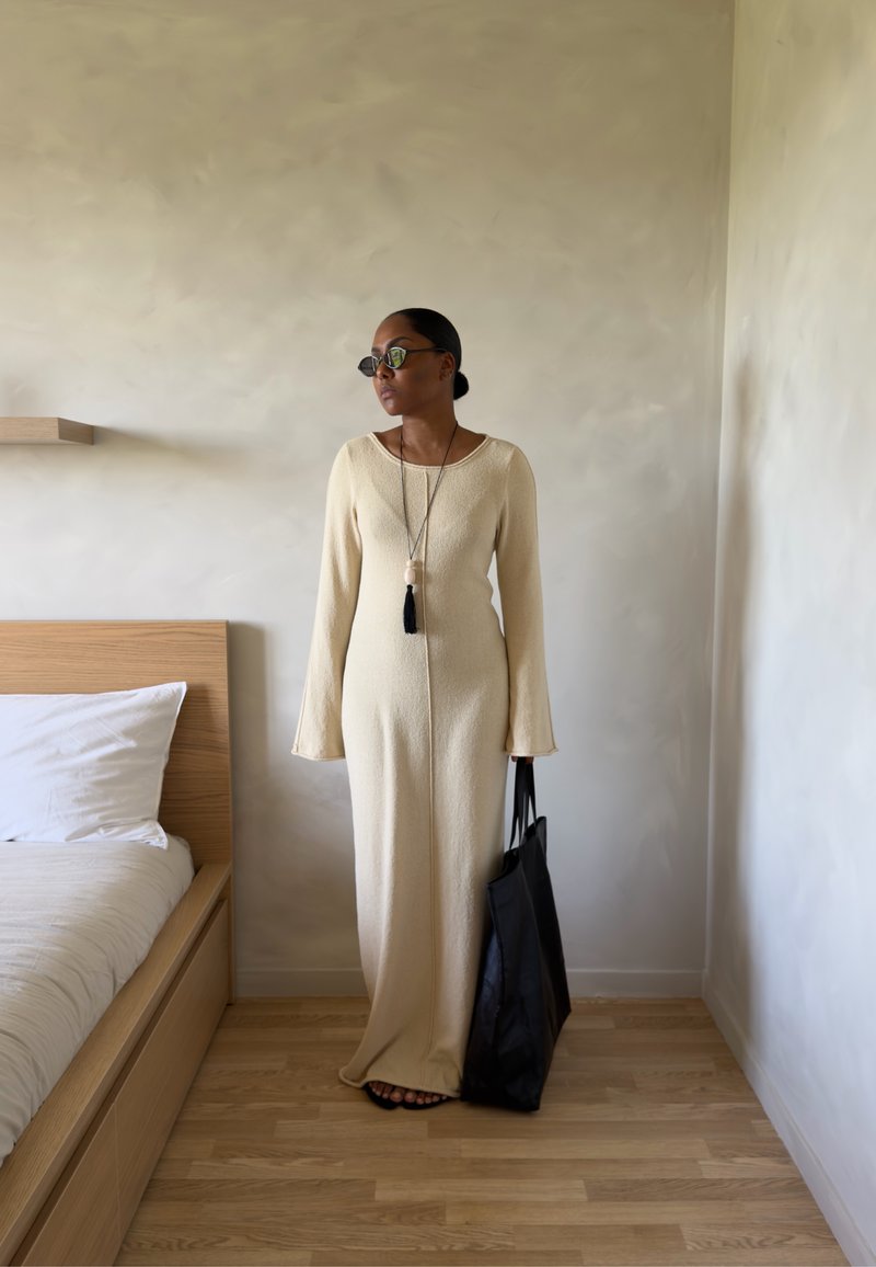 Femme en robe longue beige et lunettes de soleil debout à côté d’un lit tenant un grand sac fourre-tout noir dans une pièce minimaliste avec un sol en bois.