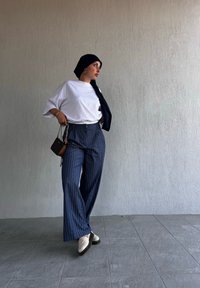 T-shirt blanc surdimensionné, pantalon large à rayures bleu marine, écharpe noire, boucles d'oreilles en or, petit sac à main noir et chaussures blanches. Fond clair.