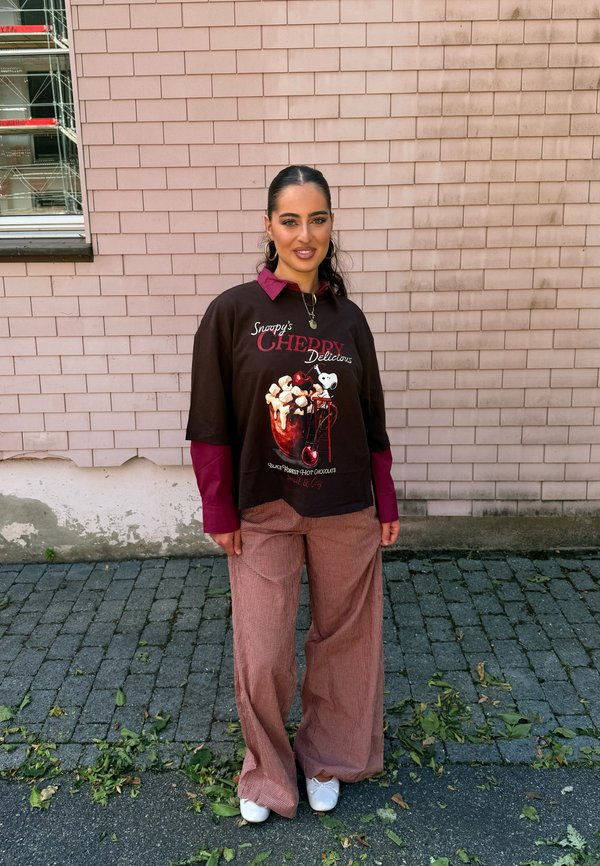 Vrouw met donker haar in een paardenstaart, gekleed in een zwarte Snoopy grafische trui over een rood shirt en wijde bruine broek, staand op een kasseienstraat.