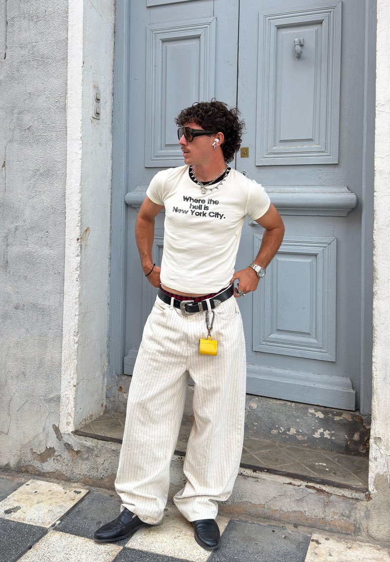 T-shirt branca com texto preto, calças largas às riscas, cinto preto com um porta-chaves, sapatos pretos, óculos de sol e auscultadores. Fundo de porta cinza claro.