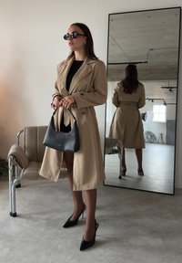Femme portant un trench-coat beige, une robe noire et des talons, tenant un sac à main noir, debout près d'une chaise et d'un miroir en pied dans une pièce moderne.