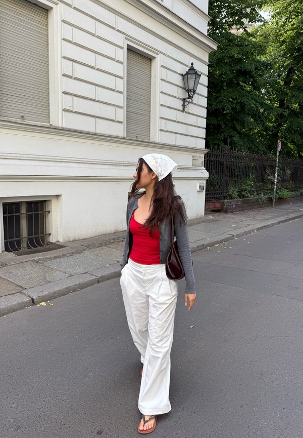 Woman wearing white headscarf, red top, gray cardigan, and white pants walking on street beside white building and green trees.