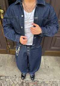 Jeans jakna in prevelikih kavbojkah v temno modri barvi, s srebrnimi okovi in dodatki iz verige. Bela srajca s črnim ročno napisanem besedilu. Črne čevlje.