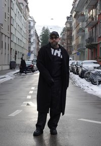 Homme portant un manteau noir, une casquette, des lunettes de soleil et des gants, marchant dans une rue de ville mouillée bordée de voitures garées et de trottoirs enneigés.