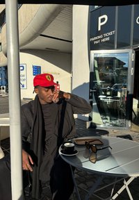 Κόκκινο καπέλο Ferrari, μαύρο μπουφάν με υφασμάτινο κασκόλ, διάφανο ποτήρι στο χέρι, φλιτζάνι μαύρου καφέ σε ασημένιο στρογγυλό τραπέζι, γυαλιά ηλίου κοντά.