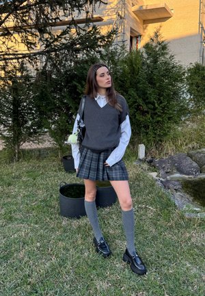 Joven mujer que lleva un chaleco de punto gris, camisa blanca, falda de cuadros, calcetas hasta la rodilla y zapatos negros, de pie sobre el césped cerca de arbustos y rocas.