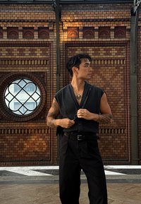 Black sleeveless vest with a deep V-neck, worn over black trousers. Circular window in the background, featuring a red brick wall with patterns.