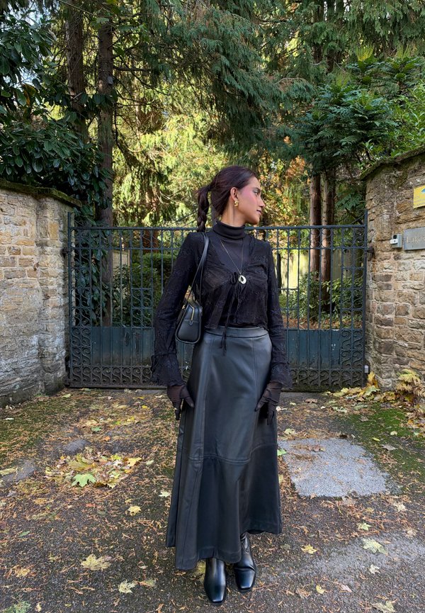 Une femme vêtue de noir se tient devant un portail en fer orné et regarde sur le côté dans un jardin verdoyant.