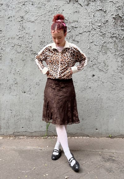 Chaqueta de polipiel con estampado de leopardo y detalles en blanco, falda marrón con flores, medias blancas con puntos, y zapatos negros con hebillas. Fondo de pared texturizada en gris.
