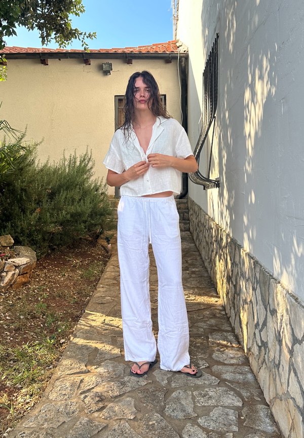 Jeune personne aux cheveux mouillés portant une chemise blanche à manches courtes, un pantalon blanc et des sandales noires, debout sur un chemin en pierre près d'une maison.