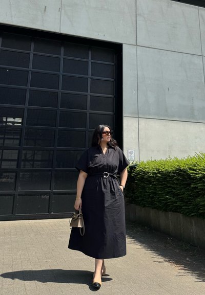 Vestido midi negro con mangas cortas y cintura ceñida, combinado con un pequeño bolso beige y unos zapatos de tacón bicolor en negro. Escenario al aire libre.