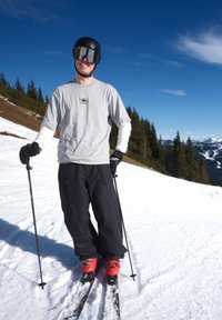Un skieur souriant portant un casque, des lunettes, une chemise grise, un pantalon noir et des bottes rouges se tient sur une pente enneigée avec des bâtons de ski, avec en arrière-plan une montagne et une forêt.