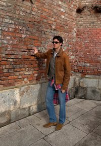 Jeune homme en veste marron, jean bleu et lunettes de soleil, debout sur un pavé en pierre, touchant un vieux mur en briques rouges.