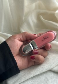 KIKO Lippenstift in einem klaren Röhrchen mit einer abgerundeten silbernen Kappe, das ein pinkes Produkt enthält, in einer Hand vor einem hellen Stoffhintergrund gehalten.