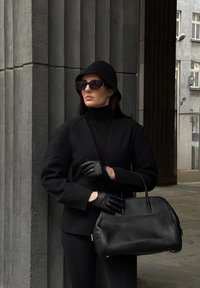 Sort struktureret blazer over en sort rullekrave, sorte handsker og en tekstureret sort håndtaske. Modellen bærer en sort hat og solbriller.