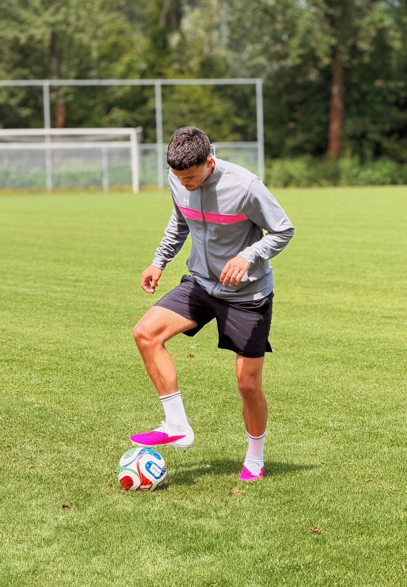 Athlète en veste grise et short noir contrôle le ballon de football avec le pied sur un terrain en herbe verte avec des poteaux de but en arrière-plan.