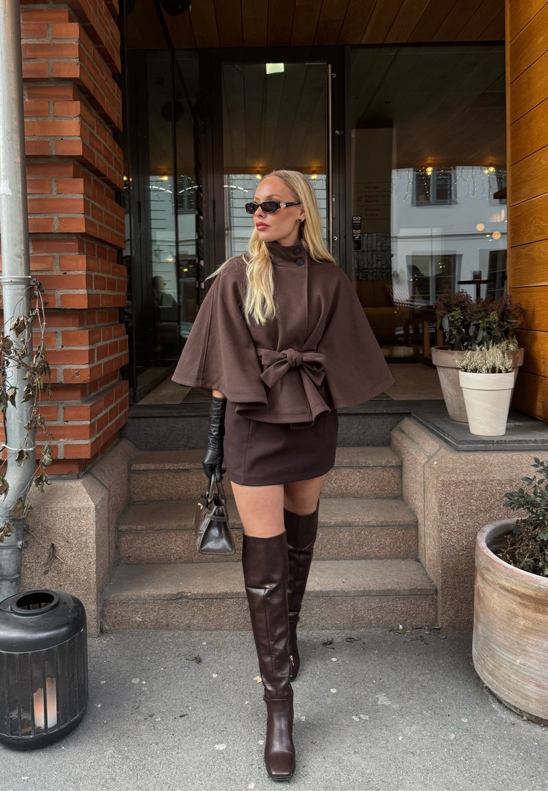 Plavokosa žena u tamnosmeđem ogrtaču s mašnom, kratkoj suknji, čizmama do koljena, crnim rukavicama i sunčanim naočalama stoji na vanjskim stepenicama pokraj biljaka.