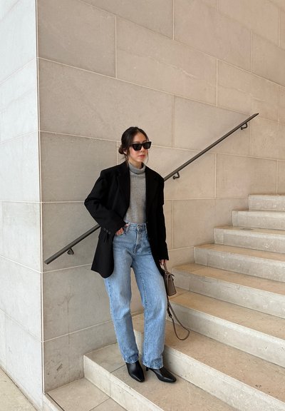 Blazer negro de corte oversize, suéter de cuello alto gris, jeans rectos de un tono azul claro, botines negros y un bolso pequeño marrón sobre una escalera de piedra clara.