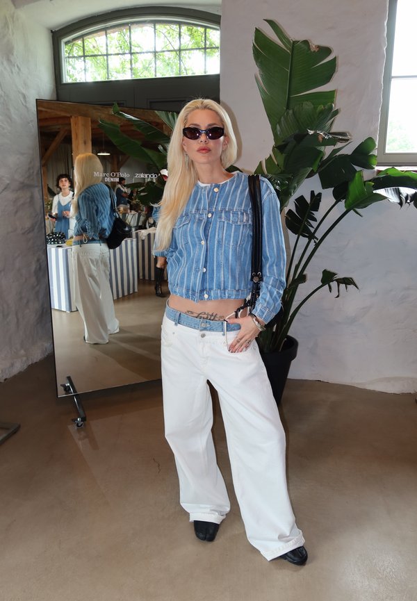 Vrouw met lang blond haar die een zonnebril, een blauw gestreept jasje en witte jeans draagt, staat binnen naast een spiegel en een grote groene plant.