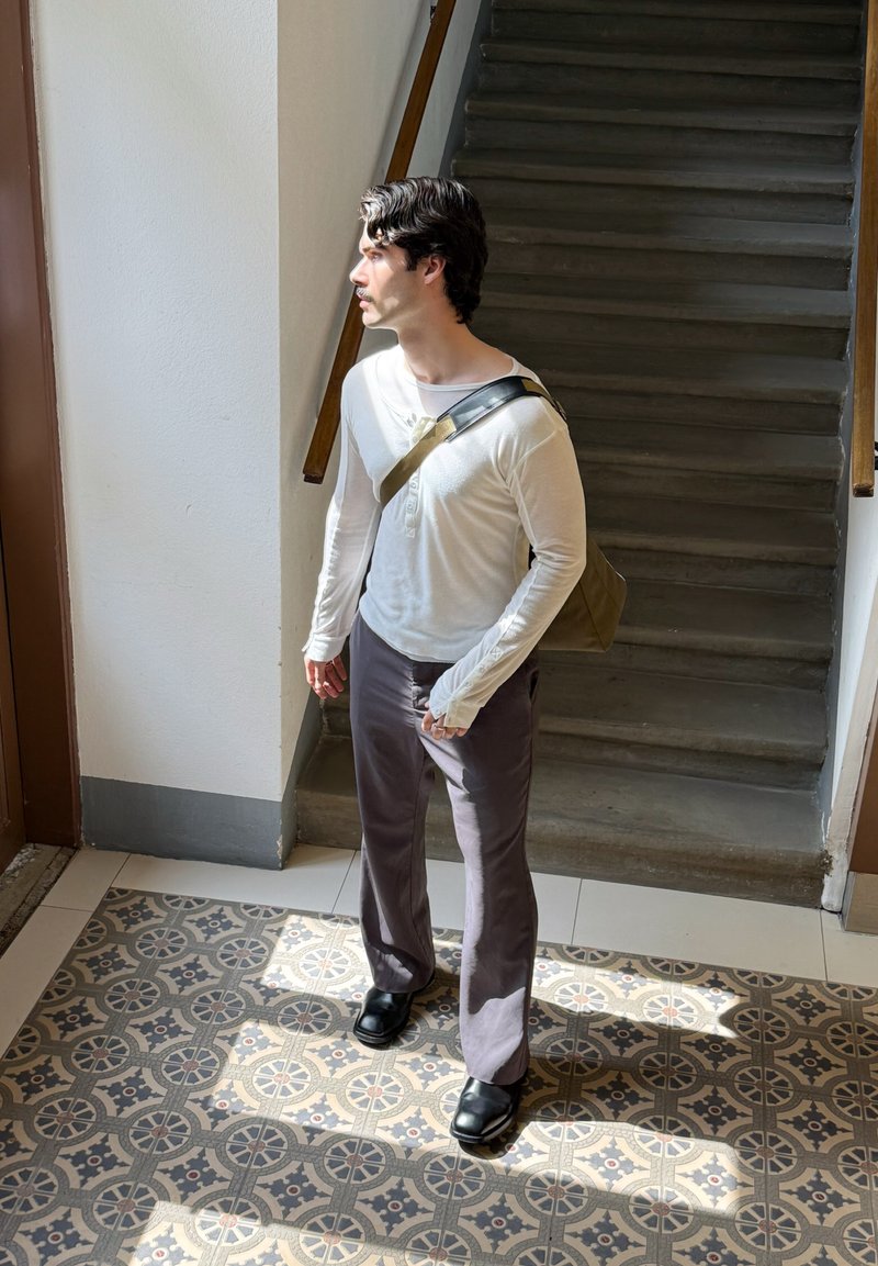 Jeune homme avec sac à bandoulière debout près d'un sol en carreaux à motifs près de l'escalier, regardant vers une fenêtre éclairée, à l'intérieur en journée.
