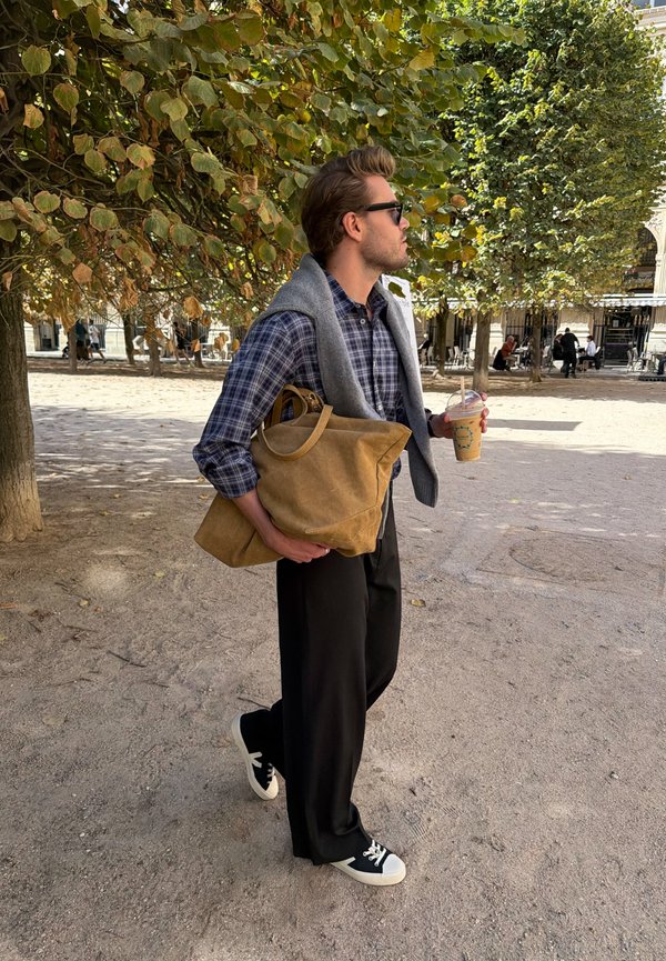 Hombre con gafas de sol y camisa de cuadros, sosteniendo una bolsa marrón claro y una bebida fría, caminando al aire libre bajo árboles frondosos en un día soleado.