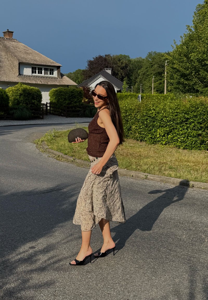 Femme portant des lunettes de soleil, un top sans manches, une jupe à motifs et des talons qui marche sur une route près de haies vertes et d'une maison au toit en chaume.