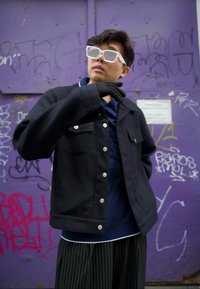 Černá bunda se stříbrnými knoflíky přes modrou košili, doplněná pruhovanými kalhotami a černými rukavicemi. Celý vzhled završují bílé úhlové sluneční brýle.