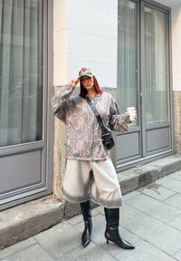 Junge Frau in einer Camouflage-Jacke, grauen weiten Shorts, schwarzen Stiefeln und einer gemusterten Mütze, die auf einem Bürgersteig in der Stadt einen Kaffeebecher hält.