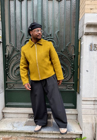 Hombre con chaqueta mostaza, pantalones anchos negros, zapatos beige y gorro negro apoyado en una ornamentada puerta metálica verde junto a escaleras de piedra.