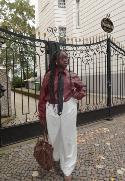 Camisa de cuero marrón con una corbata negra, combinada con pantalones anchos blancos. Bolso marrón, enmarcado por una reja de hierro decorativa.
