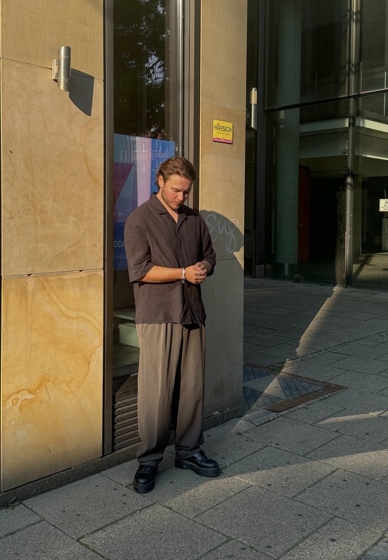 Μαύρο κοντομάνικο πουκάμισο, μπεζ φαρδιά παντελόνια και μαύρες πλατφόρμες. Στηρίζει το σώμα του σε ένα πέτρινο τοίχο, με τη σκιά του να πέφτει στο έδαφος.