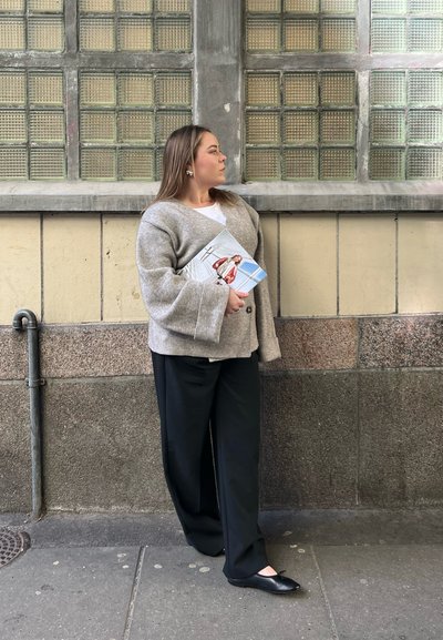 Abrigo de lana gris de gran tamaño con mangas anchas, combinado con pantalones de pierna ancha negros y bailarinas negras, sosteniendo una revista contra una pared texturizada.