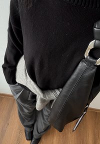 Personne portant un pull noir, un pantalon gris avec un pull gris noué autour de la taille, des bottes noires et portant un sac en cuir noir.