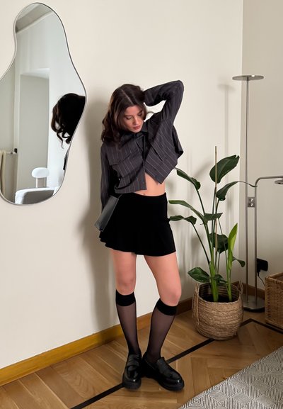 Camiseta negra corta con rayas blancas, falda negra acampanada, calcetas negras hasta la rodilla y zapatos negros gruesos. Ambiente interior con vegetación.