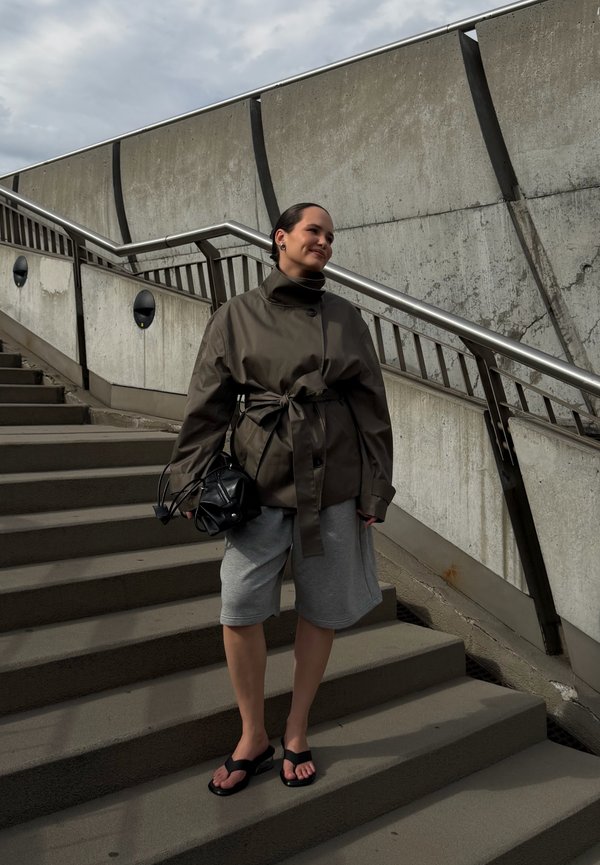 Femme en veste marron oversize et short gris debout sur des marches en béton extérieures à côté de rampes métalliques sous un ciel nuageux.