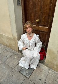 Fehér, hímzett ruha hosszú fodros ujjakban, csipke részletekkel és lebegő szoknyával. Piros táska a közelben. Fából készült ajtó a háttérben.