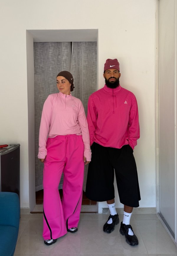 Dos personas de pie en el interior vistiendo camisetas deportivas rosas, la mujer con pantalones anchos rosas y pañuelo en la cabeza, el hombre con pantalones cortos anchos negros y gorra.