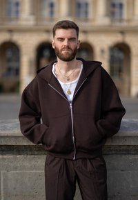 Brauner Reißverschluss-Hoodie mit großer Kapuze, getragen über einem weißen Shirt, kombiniert mit passenden braunen Hosen. Hat eine lockere Passform und seitliche Taschen.